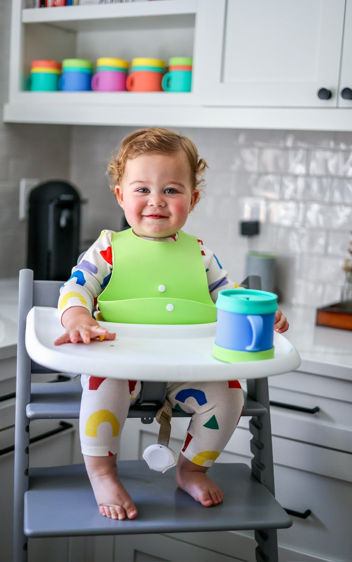 Smiling toddler in a high chair wearing a green Roll & Snap Bib with a Morepeas Snack Cup