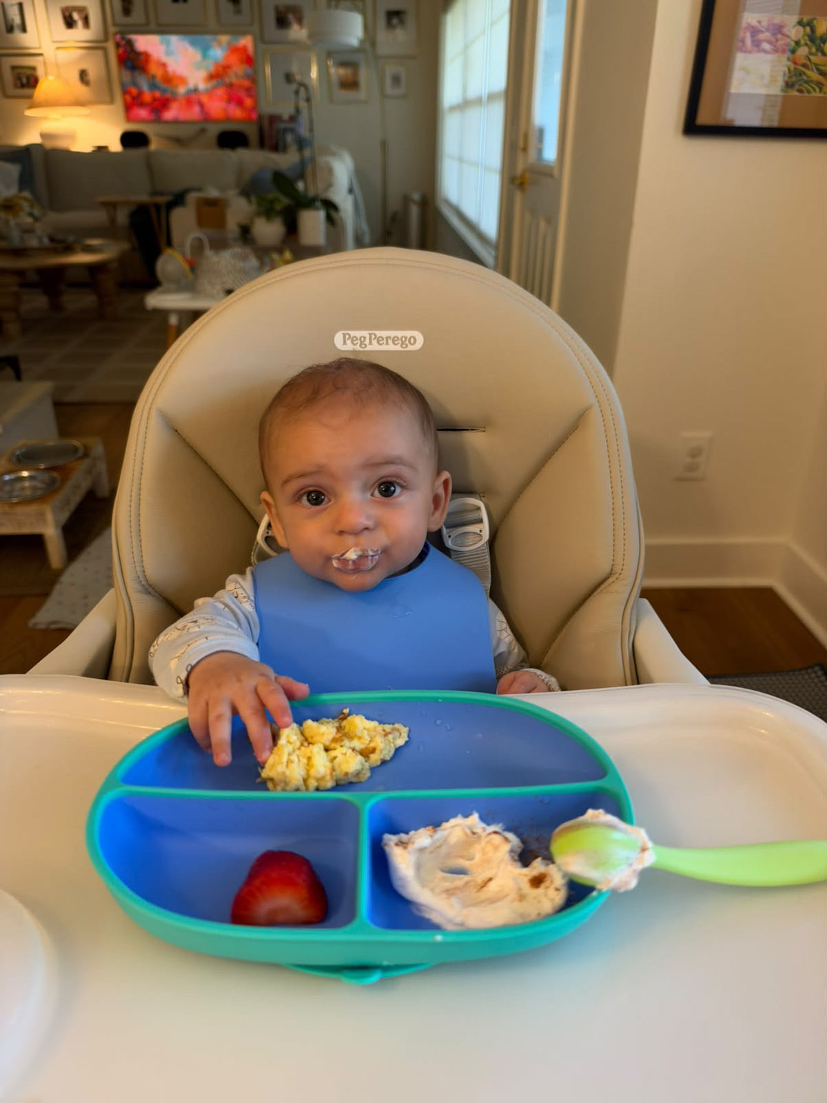 Baby in a high chair eating from a Morepeas suction plate, wearing a blue Roll & Snap Bib, with a Baby to Tot spoon